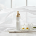Vaporisateur d'ambiance blanc nacré et doré sur marbre blanc, entouré d'orchidées blanches et de tissu fluide